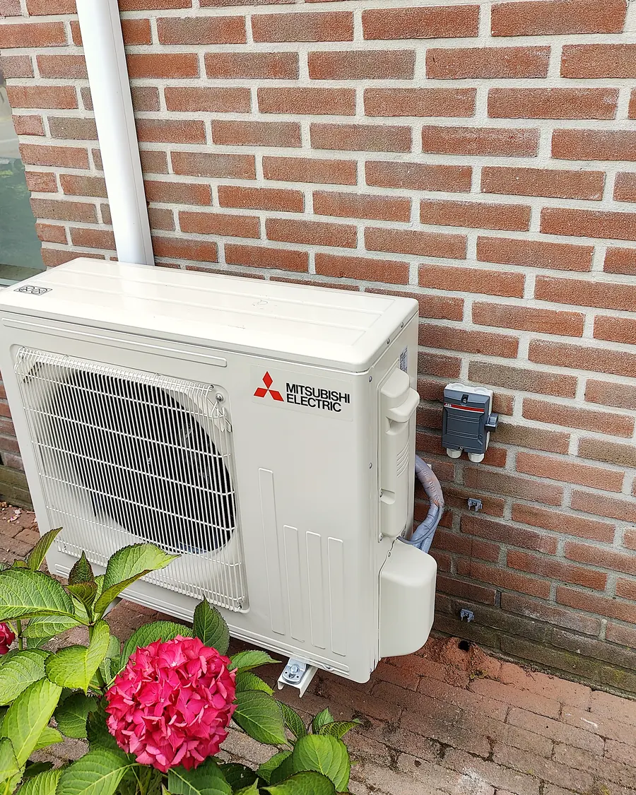 Buitenunit van een airco aan een bakstenen gevel, met een bloeiende hortensia ervoor