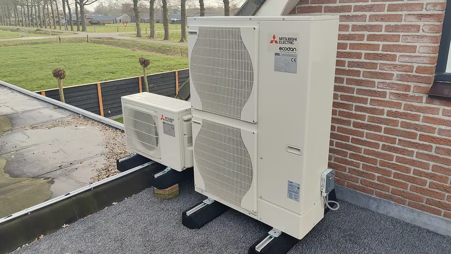 Twee buitenunits van een lucht-waterwarmtepomp op een plat dak tegen de gevel, met uitzicht over het weiland